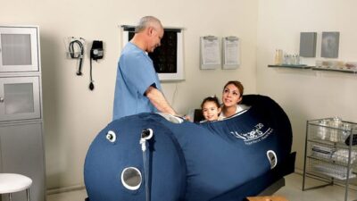 A doctor attends to a mother and child seated inside a Zuegma OxyHealth Hyperbaric Chamber in a clinical setting.