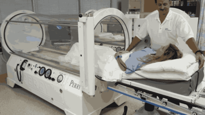 A patient lying on a bed is being guided into a Perry brand hyperbaric chamber by a medical professional — perry hyperbaric chamber.